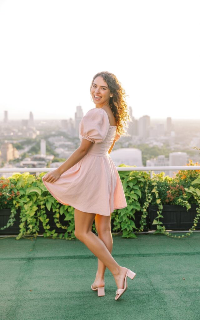 The model stands on a rooftop garden at golden hour wearing a pastel pink puff-sleeve dress with a fitted waist and flared skirt. Her hair is styled in loose curls cascading over one shoulder. The warm sunlight enhances her skin tone and adds a soft glow. She wears block heels and no other accessories. Her pose is playful, one hand holding her skirt slightly as she looks back with a bright smile.