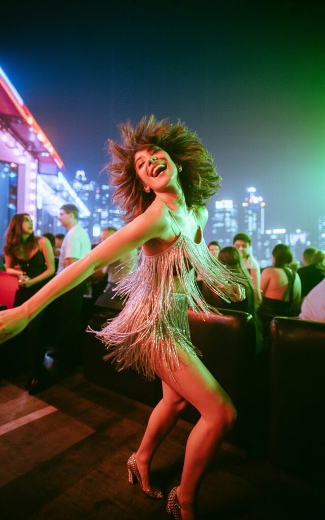 White-skinned model with curly brown hair, wearing a silver fringe mini dress with metallic heels. Outdoor rooftop club scene at night, neon lights glowing. She’s mid-dance, fringe flying, head tilted back in laughter.