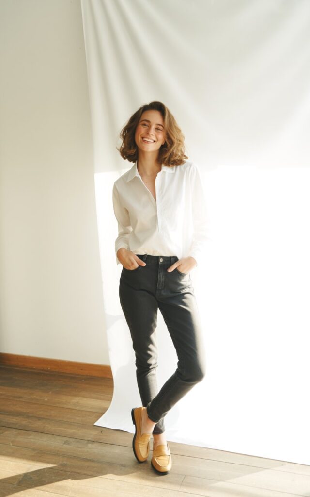 White-skinned model with natural brunette waves, wearing dark wash skinny jeans, crisp white tucked button-up, and tan loafers. Captured in a sunlit modern studio with wooden flooring. She’s standing tall, hands tucked in pockets, smiling naturally, giving timeless simplicity.