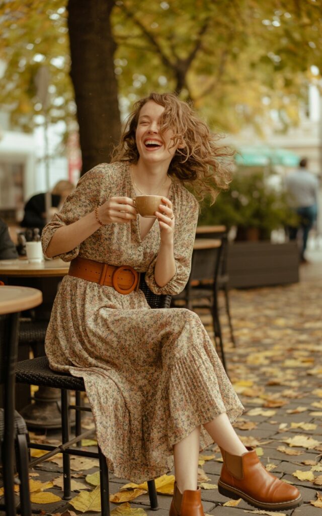 At an outdoor café surrounded by autumn leaves, a white-skinned woman with light brown curls wears a flowy floral midi dress cinched with a wide belt and ankle boots. Natural daylight, soft breeze moving her hair. She stands holding a coffee cup, laughing mid-candid, radiating cozy boho charm.