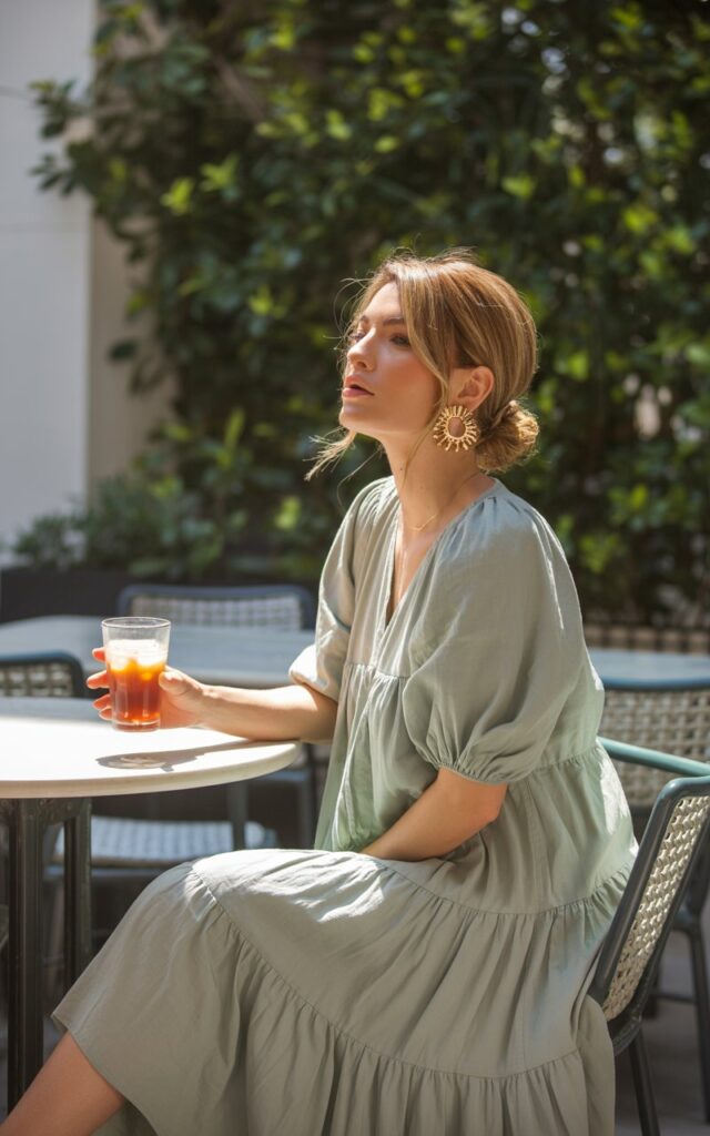Captured in a sunlit café patio scene. The model sits elegantly with a drink, wearing a sage green tiered midi dress and chunky gold earrings. Her hair is styled in a low messy bun with soft strands framing her face. Natural daylight filters through greenery, highlighting the texture of her dress.