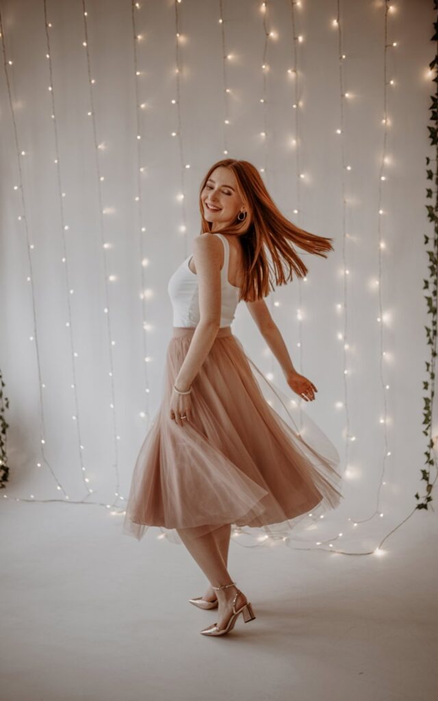 Dreamy studio backdrop with fairy lights. Model in a blush tulle midi skirt paired with a fitted white tank top, delicate heels, and subtle jewelry. Spinning gently, dreamy smile, ethereal vibe.