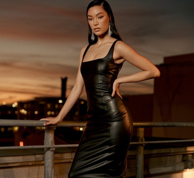 Evening city rooftop setting. Model with sleek black hair wears a fitted black leather midi dress, heeled boots, and statement earrings. Warm city lights and sunset tones in the background. She poses with one hand on the railing, expression confident and sultry.