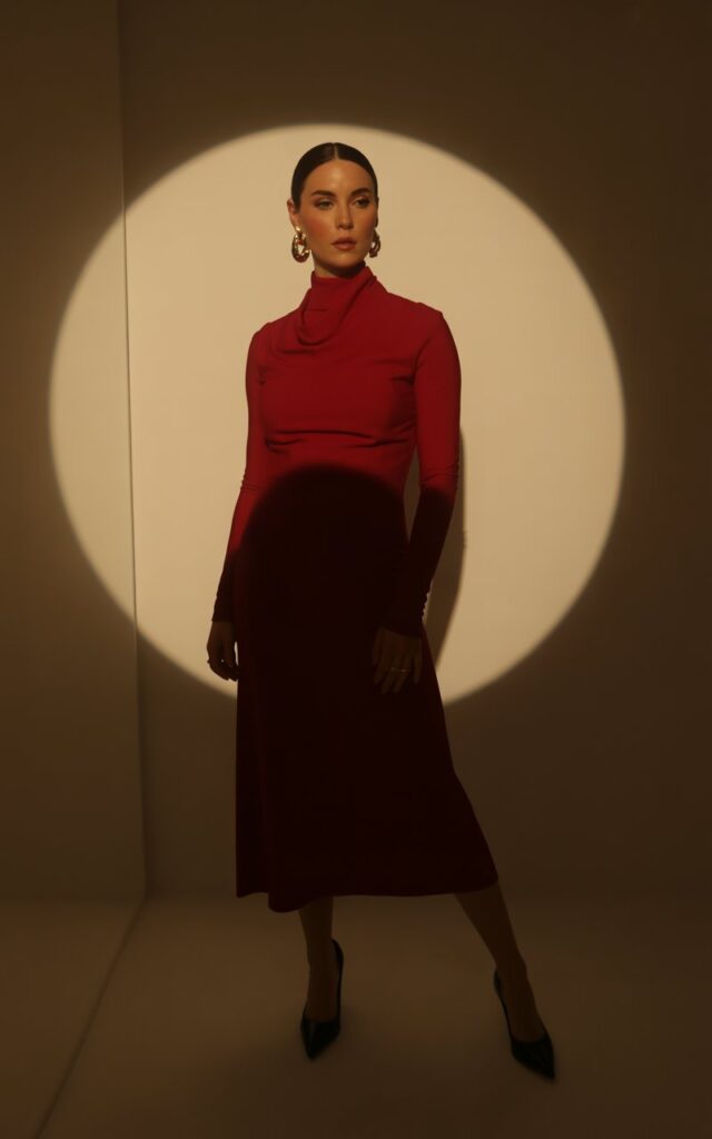 Full-body photo of a white-skinned woman with slicked-back hair wearing a crimson high-neck midi dress and bold gold earrings. She’s standing under soft indoor spotlight lighting with a minimalist beige background. Elegant stance with one leg slightly forward, hands relaxed at her sides, refined expression.
