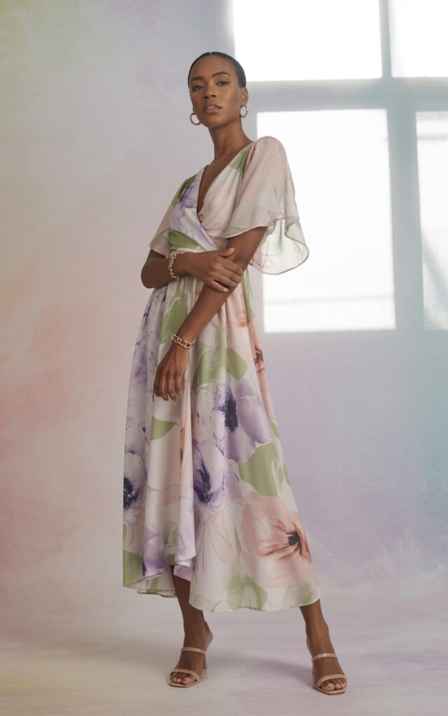 Full-body view in a pastel studio backdrop. The model wears a watercolor floral wrap dress, nude sandals, and minimal jewelry. Soft natural lighting highlights the print’s delicacy. She’s posed elegantly with one hand resting on her hip.