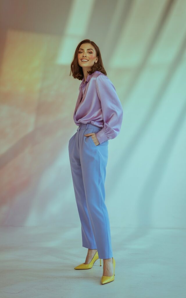 Full-body view in a pastel-toned studio with soft lighting. The model wears a lavender blouse, baby-blue trousers, and pale-yellow heels. Her makeup is dewy and minimal, and she’s smiling confidently with one hand in her pocket. The pastel background complements the outfit.