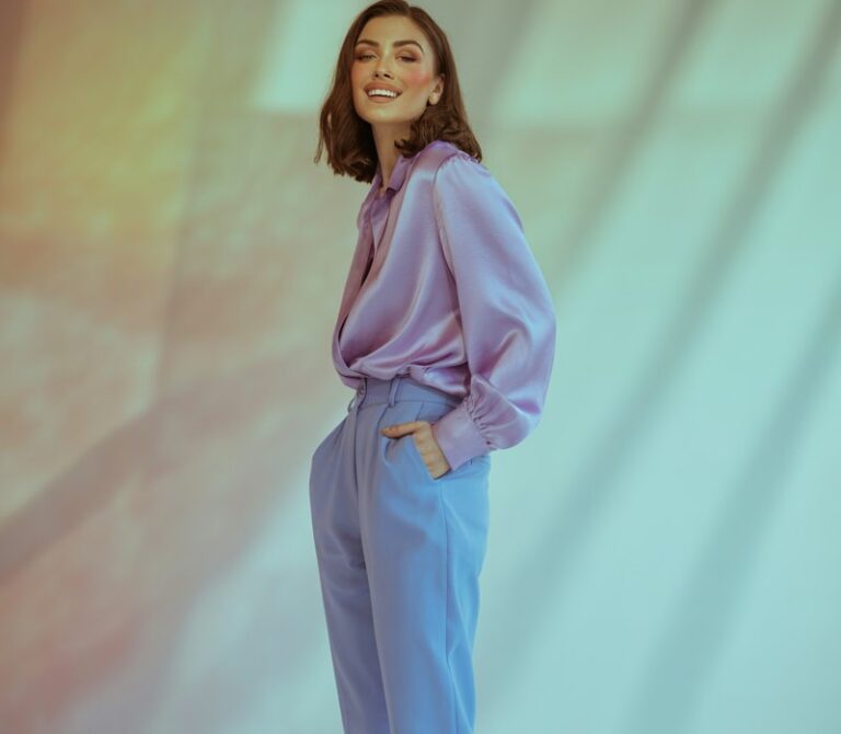 Full-body view in a pastel-toned studio with soft lighting. The model wears a lavender blouse, baby-blue trousers, and pale-yellow heels. Her makeup is dewy and minimal, and she’s smiling confidently with one hand in her pocket. The pastel background complements the outfit.