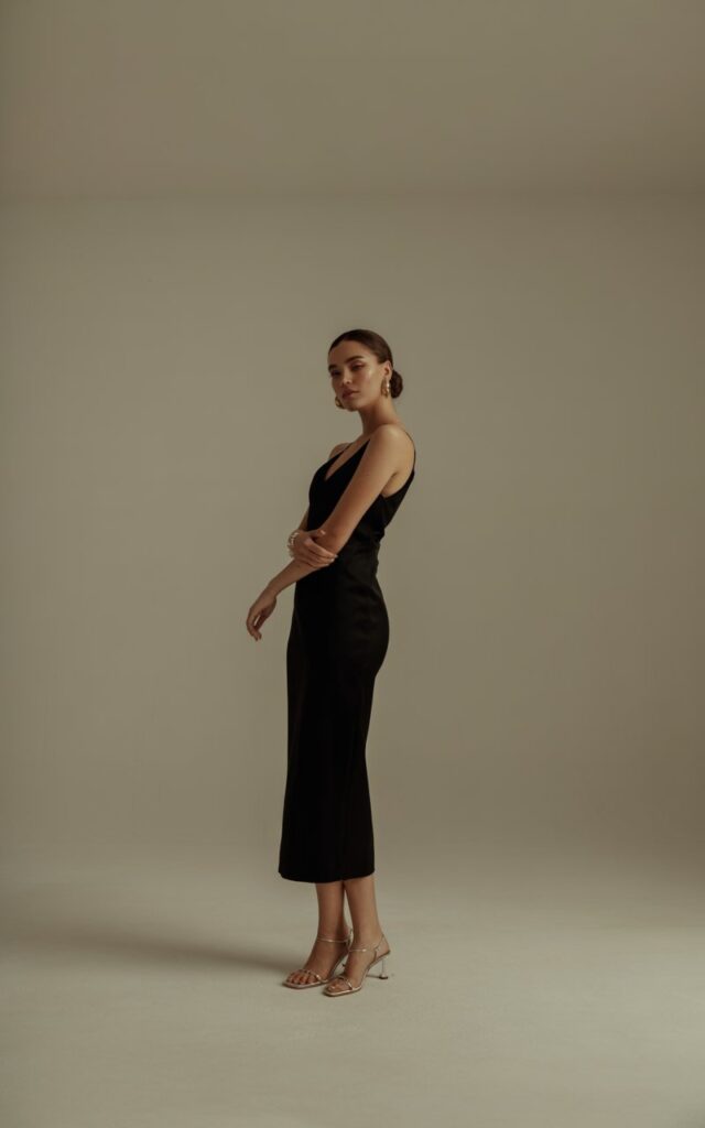 Indoor editorial shot against a neutral beige backdrop. Model stands tall in a sleek black midi dress with strappy sandals, dainty gold hoops, and hair in a low bun. Soft directional light adds gentle contrast. Pose slight turn of the body, elegant and confident expression.