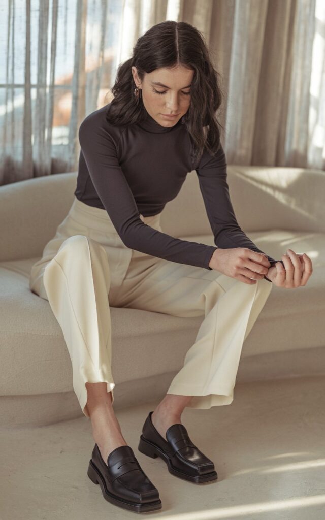 Indoor lifestyle image of a white-skinned woman with dark wavy hair wearing sleek black loafers, cream trousers, and a tucked-in turtleneck. Cozy, minimalist home setting with sunlight filtering through sheer curtains. She’s adjusting her cufflinks, expression calm and thoughtful.