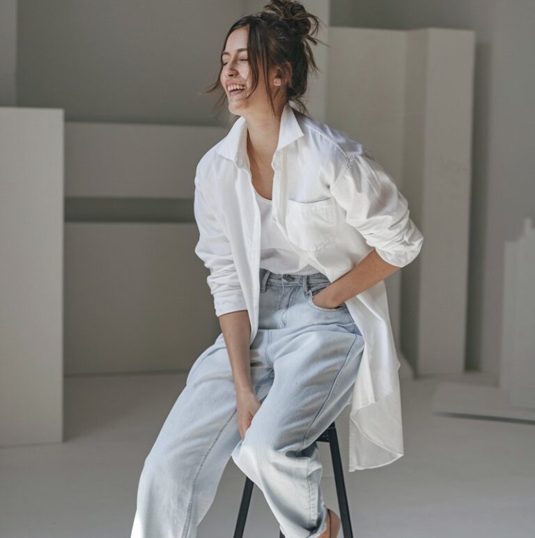 Minimal studio backdrop with white tones. White-skinned model with messy bun wears a crisp white shirt, light blue relaxed jeans, and nude heels. Lighting is bright natural daylight. She sits on a stool mid-laugh — timeless and effortless.