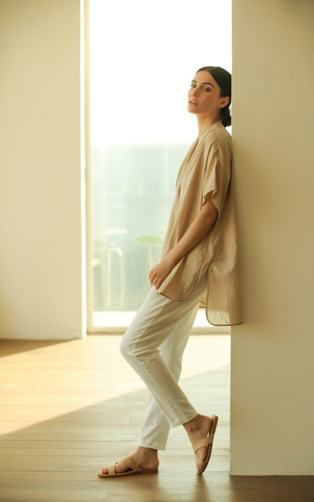 Minimalist indoor setting with soft natural light. The model stands near a large window wearing a beige oversized tunic with white slim cotton pants and leather sandals. Her hair is tucked behind her ears, makeup fresh and simple. She leans slightly on the wall, exuding quiet elegance and ease.