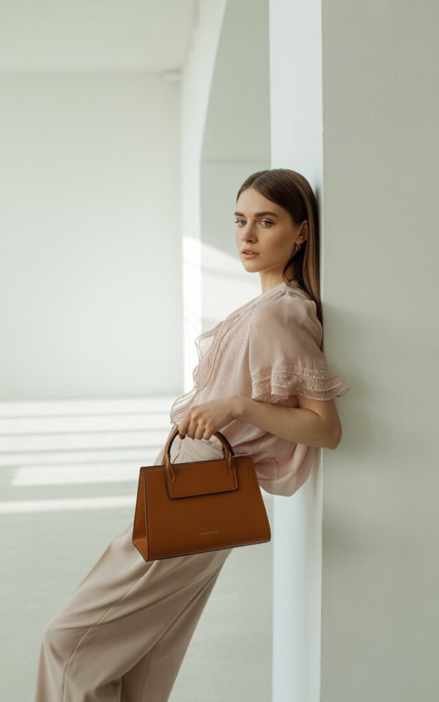 Model leaning against a white studio wall wearing a ruffled blush blouse, tailored beige trousers, and a structured leather handbag. Soft daylight filters through large windows. Her hair is sleek and parted, giving balance to the playful ruffles.
