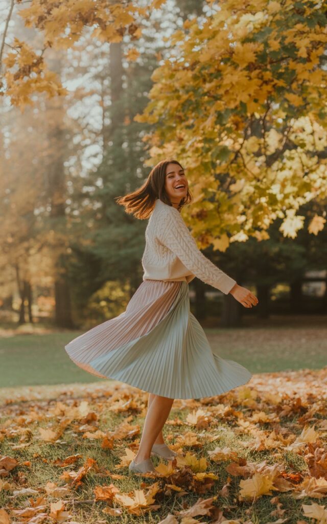 Model mid-twirl in a park during autumn. She wears a pastel pleated midi skirt and a tucked-in cream sweater. The leaves create a golden backdrop. She’s laughing, with her skirt flowing around her and natural light catching the movement.