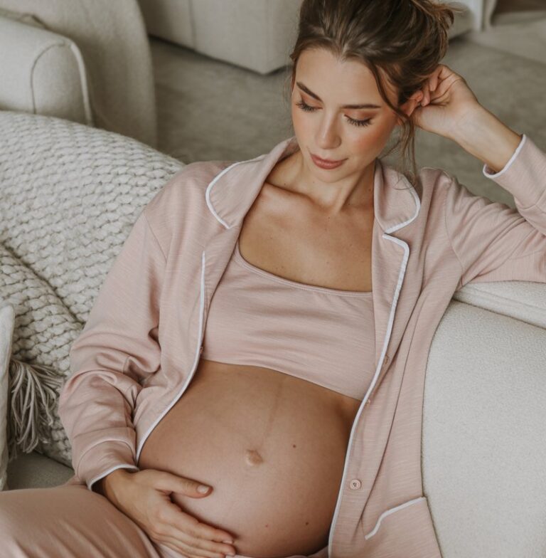 Model pregnant women in a blush two-piece lounge set, lounging on a plush sofa. Soft indoor lighting with a cozy blanket nearby. Minimal makeup, hair in a messy bun, peaceful expression.