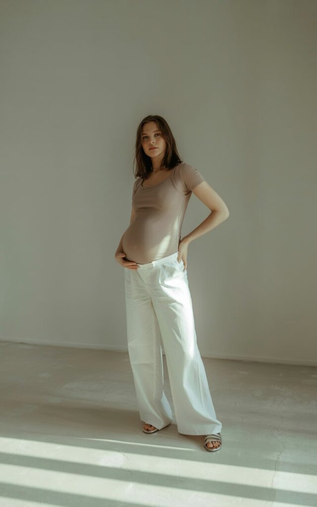 Model pregnant women in a fitted beige maternity bodysuit tucked into flowy white wide-leg trousers. Minimalist studio with diffused light. Confident stance, hands on hips, clean and editorial feel.