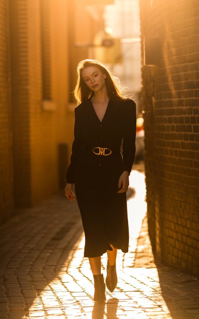 Outdoor brick alleyway with golden sunlight. White-skinned model with light freckles wears a black midi dress, bold gold-buckled belt, and ankle boots. Lighting is golden hour. She looks over her shoulder mid-step, effortlessly confident.