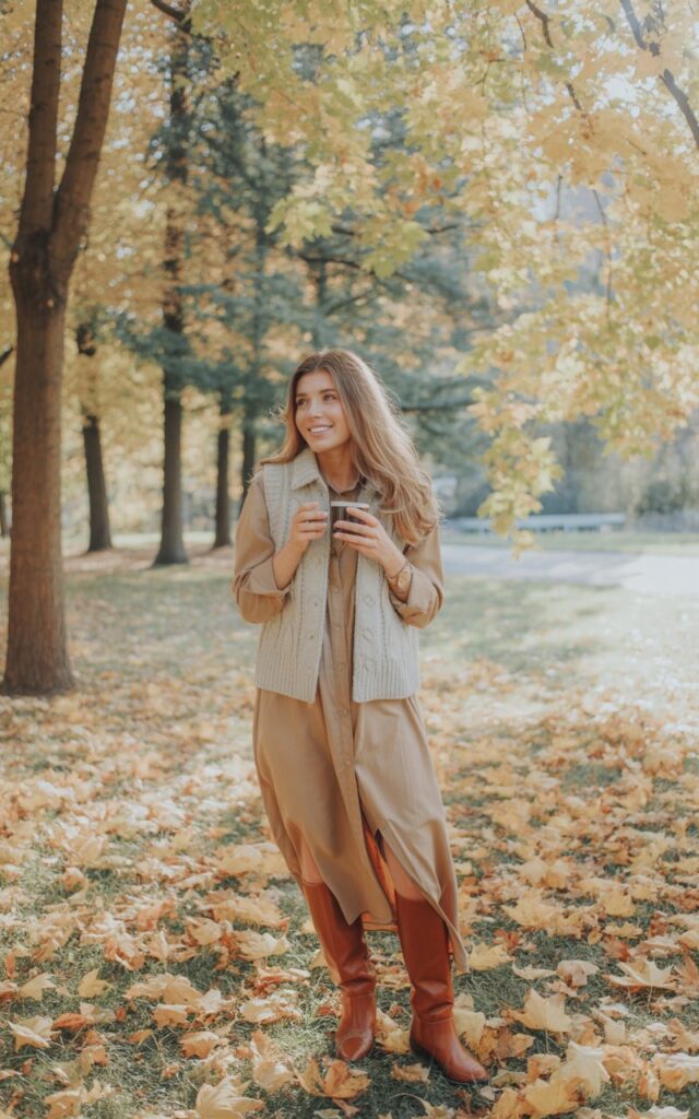 Outdoor fall park scene with golden leaves. The model wears a cream knit vest layered over a tan maxi shirt dress with knee-high boots. Her light brown hair flows naturally, and she holds a hot drink with a gentle smile. Natural daylight creates soft, diffused warmth.