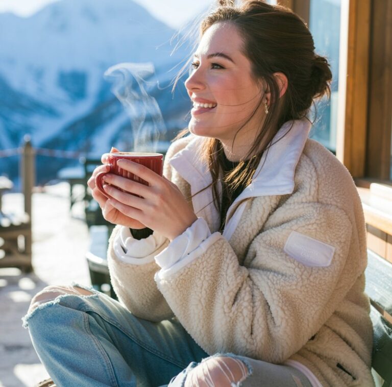 Shot in a snowy mountain café patio under bright daylight. She’s wearing a cream sherpa jacket, distressed blue jeans, and lace-up boots. A hot cocoa steams in her hands, and her cheeks are flushed from the cold. She looks playful and relaxed.