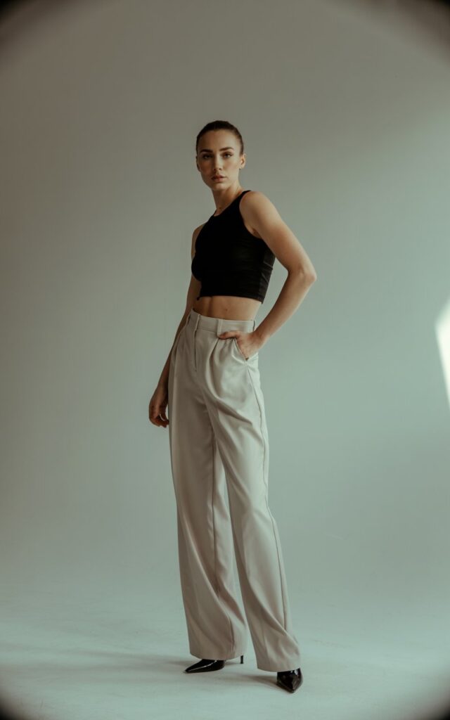 Studio portrait of a tall, fit woman in a minimalist setting. She wears a black cropped top with beige high-waisted wide-leg trousers and black heels. Soft diffused lighting enhances her sharp features. She’s standing with one hand on her hip, exuding modern sophistication.
