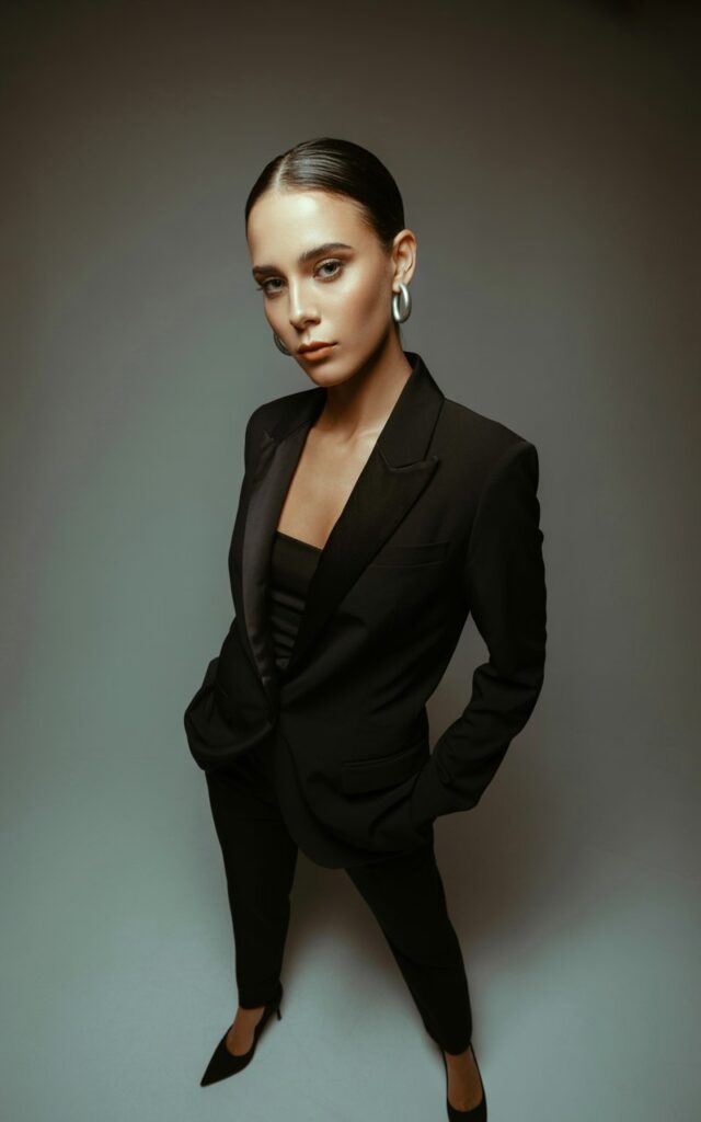 Studio portrait with dramatic soft lighting. The model wears a sleek black tailored blazer, black trousers, and black stiletto heels. Minimalist jewelry adds polish. She stands tall with hands in pockets, hair slicked back into a low bun, giving a sharp, commanding expression.