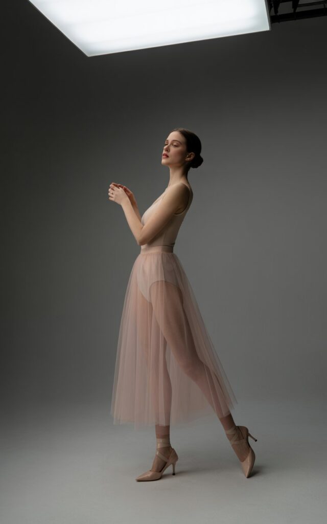 Studio setting with dramatic lighting. Model in a soft pink sheer tulle midi skirt layered over a nude fitted bodysuit, delicate heels. Hair sleek in a bun, elegant pose with arms lifted slightly, ballet-inspired.
