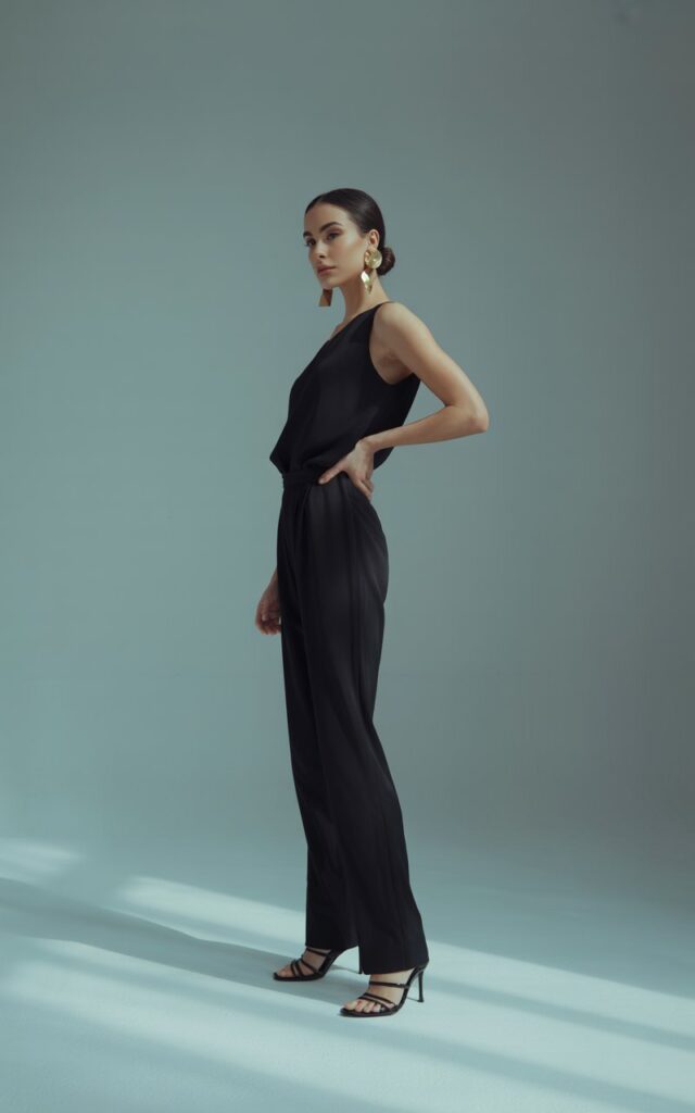 Studio setup with a soft gray backdrop. The model stands confidently, wearing a sleek black minimalist jumpsuit with bold gold earrings. She pairs it with strappy heels and a low bun. Lighting is soft and dramatic, focusing on her posture and accessories. The vibe is refined, clean, and editorial.