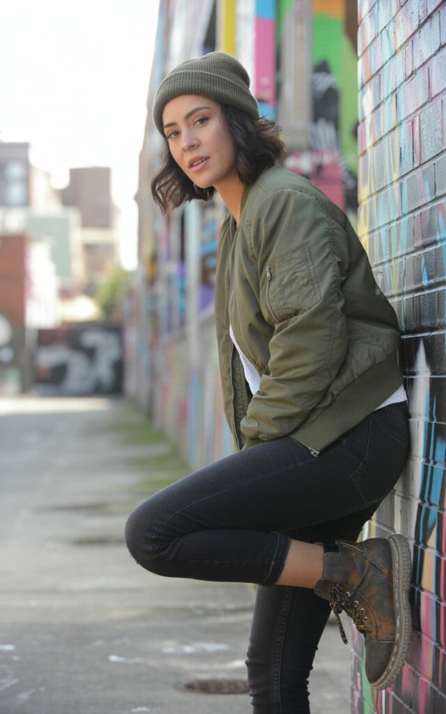 Urban graffiti alley under natural daylight. She wears an olive bomber jacket, black skinny jeans, and chunky combat boots. Her gray beanie sits low over tousled hair. She’s posing casually against a wall, one knee bent, expression confident and cool.