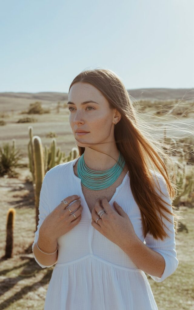 White-skinned brunette woman with long straight hair, wearing a simple white cotton dress, turquoise statement necklace and rings. Shot in a sunlit desert backdrop with cactus plants. Natural daylight, clean composition. She’s standing gracefully with calm confidence, wind lifting her hair slightly.