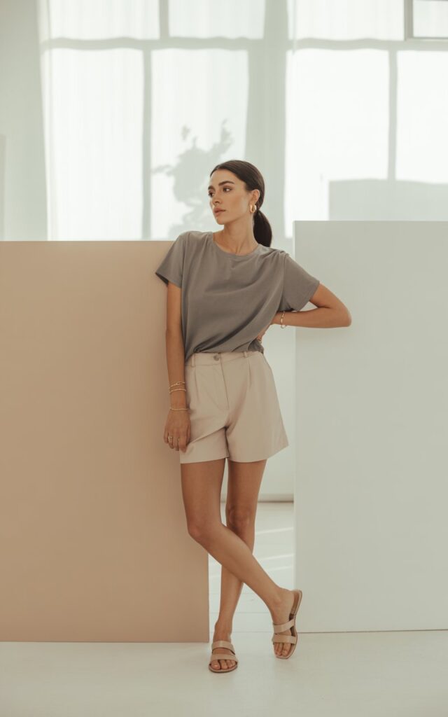 A minimalist studio with neutral tones. The woman stands with one hand in her pocket wearing a gray boxy tee tucked into beige tailored shorts, with simple sandals and delicate jewelry. Her hair is a low ponytail, makeup fresh and soft. The light is natural and clean.