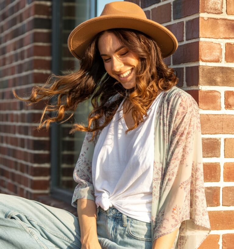 Full-body fashion photo of a white-skinned woman with chestnut waves, leaning against a brick wall in golden afternoon light. She wears a printed kimono over a plain white tee, light-wash jeans, and boho sandals. A wide-brim hat completes the look. Her playful smile feels candid and relaxed.