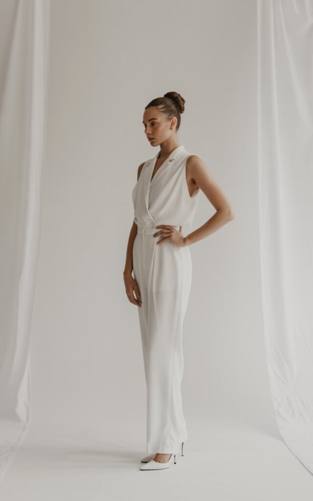 Full-body fashion shot of a white-skinned female model with a sleek high bun, wearing a fitted monochrome white jumpsuit and minimalist heels. Indoor studio with clean white walls and soft diffused lighting. She poses with one hand on her hip, expression calm and elevated. Subtle shadows and true-to-life fabric folds.