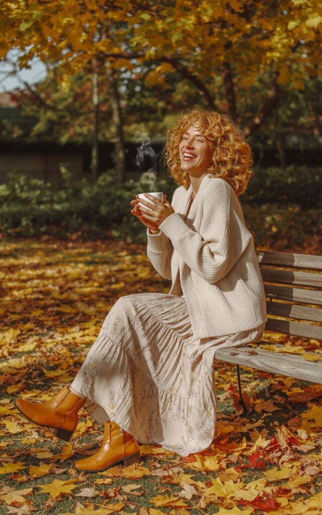 Full-body image of a white-skinned woman with curly blonde hair, sitting on a park bench surrounded by autumn leaves. She wears a chunky knit cardigan over a flowy printed maxi skirt and ankle boots. Natural daylight filters through trees as she laughs softly, holding a coffee cup.