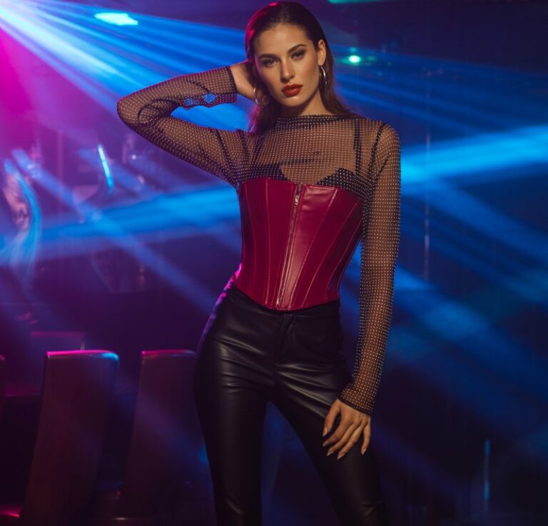 Full-body photo in a moody club-like setting. Model wears a sheer black mesh long-sleeve over a red corset, paired with leather pants and stiletto boots. Colored LED light accents (pink and blue). She stands confidently with one hand in hair, gaze direct, expression bold and alluring.
