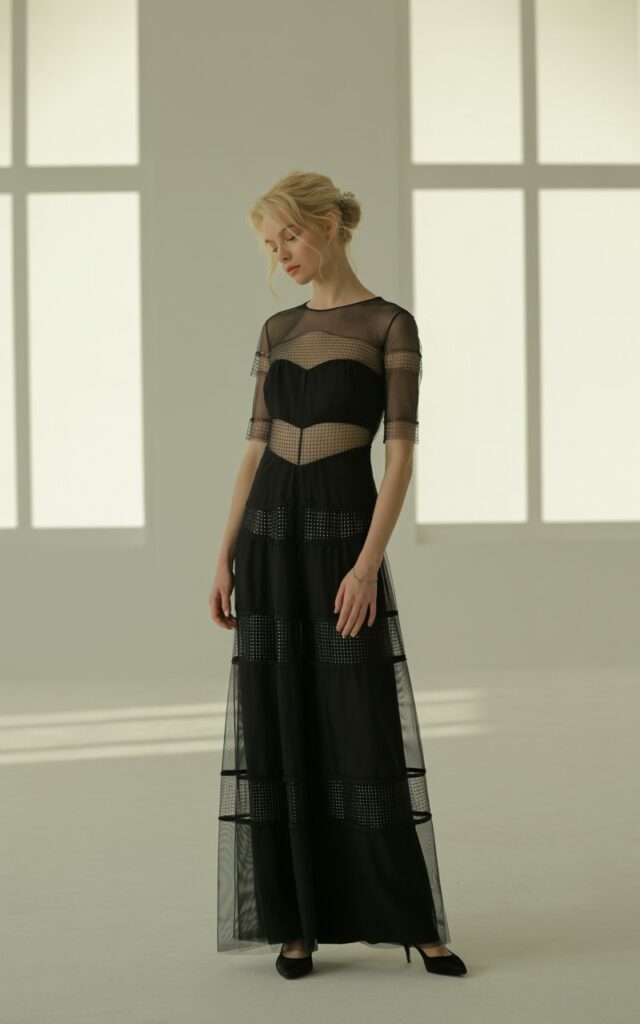 Full-body photo of a fair-skinned model in a black gown featuring illusion mesh panels placed strategically, paired with simple heels. Set inside a softly lit studio with window light. Her hair is styled in a messy bun for contrast. She stands in a poised, elegant posture with a soft expression. Mesh textures and natural skin realism emphasized.