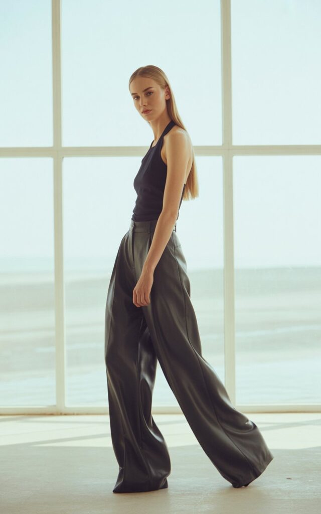 Full-body photo of a white-skinned model with sleek straight hair, wearing high-waisted wide-leg trousers and a black halter top. Bright airy studio with large windows and natural daylight. She stands in a tall elongated pose, expression composed. Flowy pant movement and skin realism emphasized.