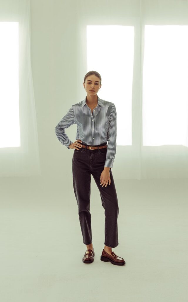 Full-body shot in a bright, minimal studio with soft window light. Model wears a blue-striped button-down tucked into straight-leg jeans, loafers, and a leather belt. Neutral background to highlight the outfit. She stands confidently with one hand on her hip. Realistic skin tone and crisp clothing texture.