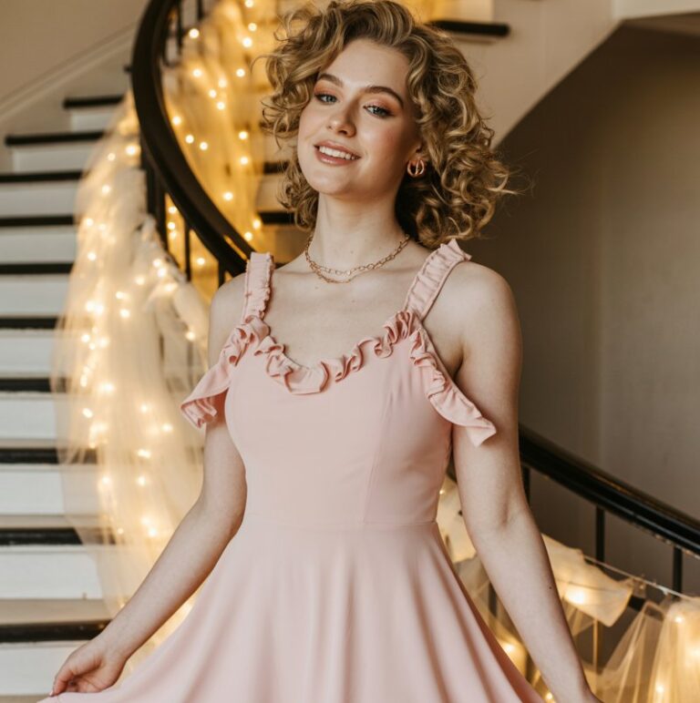 Full-body shot of a white-skinned female wearing a pastel ruffle-trim hoco dress, soft curls, and delicate jewelry. She stands near a decorated indoor staircase wrapped with tulle and fairy lights. Soft warm indoor lighting enhances the romantic vibe. She gently lifts the ruffled hem while smiling to the side.
