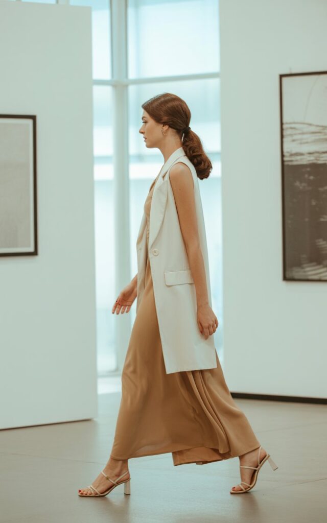 Full-body shot of a white-skinned woman with long chestnut hair in a low ponytail, wearing a white longline vest over a tan column dress with strappy sandals. Captured in a modern art gallery with bright diffused natural light. She walks gracefully, dress flowing slightly. Elegant, serene expression.