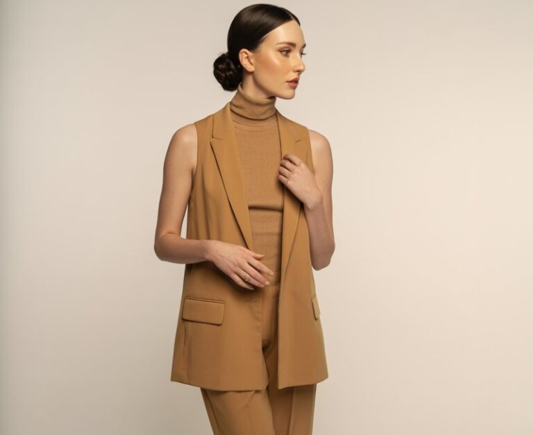 Full-body shot of a white-skinned woman with sleek low bun, wearing a camel-tone vest layered over a matching turtleneck and trousers, styled with loafers. Indoor studio with soft warm light. She stands with one hand slightly raised, as if adjusting her vest. Minimal, elegant expression.