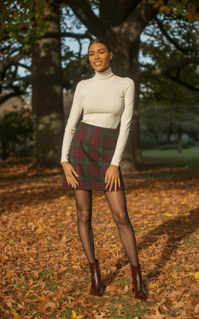 Full-body view of a model in a fitted turtleneck sweater tucked into a plaid mini skirt, black tights, and ankle boots. Autumn park setting with soft filtered sunlight. Hair sleek and straight, warm-toned makeup. Hands lightly on hips, slight tilt of head, confident smile. Natural editorial realism, textures of knit and skirt visible.