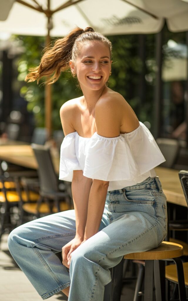 Full-body view of a white-skinned female model with a bouncy ponytail, wearing high-waisted light-wash jeans and a white off-shoulder ruffle top. Shot on a sunny café patio in natural daylight. She sits casually on a stool, leaning slightly forward with a playful smile. Realistic denim texture, soft skin glow, minimal makeup.