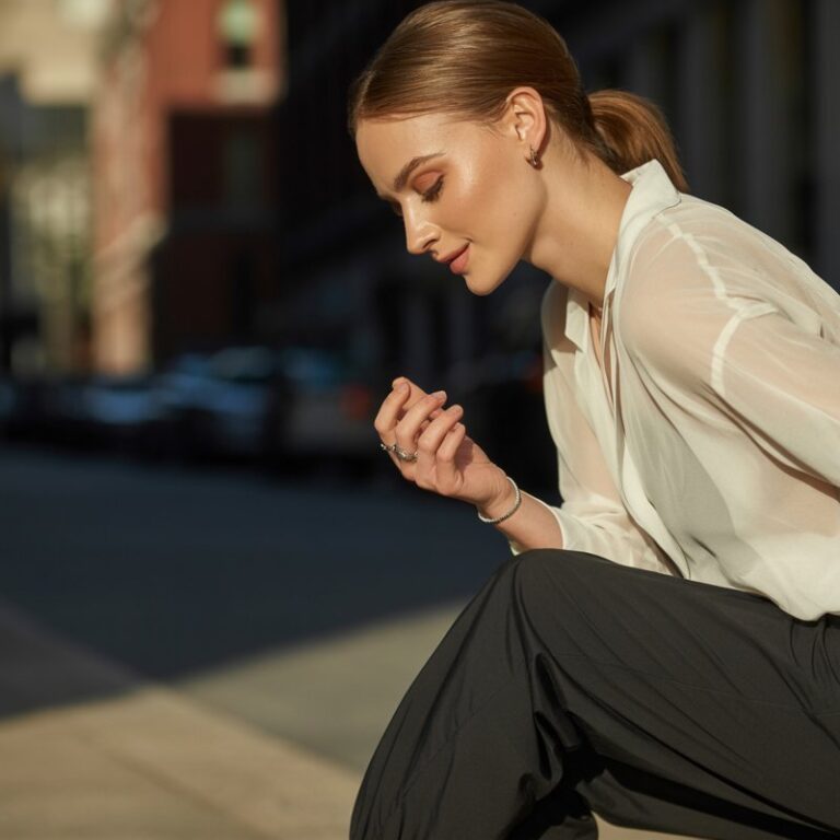 Full-body view of a white-skinned model wearing metallic silver flats with black joggers and cream blouse. Urban street with warm daylight. Hair in a low ponytail, subtle rings, soft natural makeup. Pose mid-step, looking down at shoes, gentle smile. Emphasize reflective surfaces, fabric textures, and realistic editorial styling.