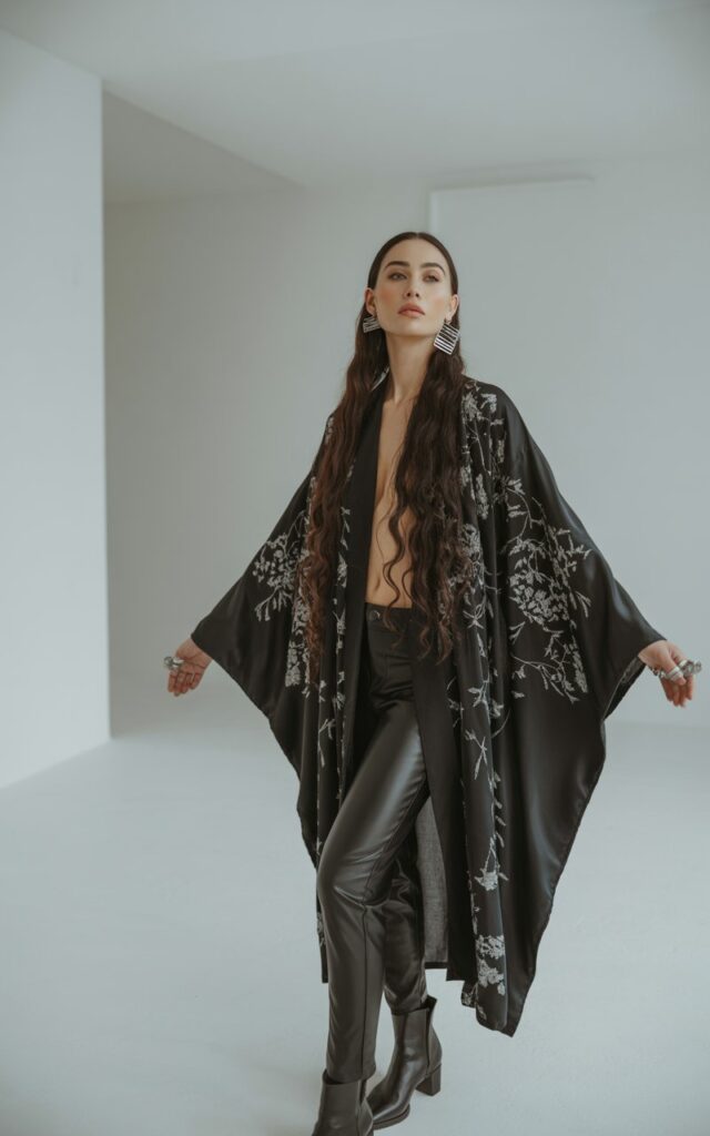 Full-body view of a white-skinned model with long dark hair, wearing a flowing black kimono with embroidered details over black leather skinny pants and black ankle boots. Accessories statement earrings, subtle rings. Pose standing in a studio with minimalistic background, arms slightly open to show kimono. Lighting soft studio lighting highlighting textures. Expression serene, head slightly tilted, natural editorial look.