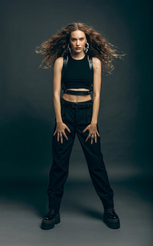 Full-body view of a white-skinned model with long wavy brunette hair, wearing a fitted black crop top, high-waisted black trousers, and a leather harness with chunky boots. Accessories silver rings, bold earrings. Pose standing confidently with hands on hips in a studio with dark background. Lighting soft studio lighting highlighting outfit contours. Expression bold and fierce, slight smirk, natural skin texture.