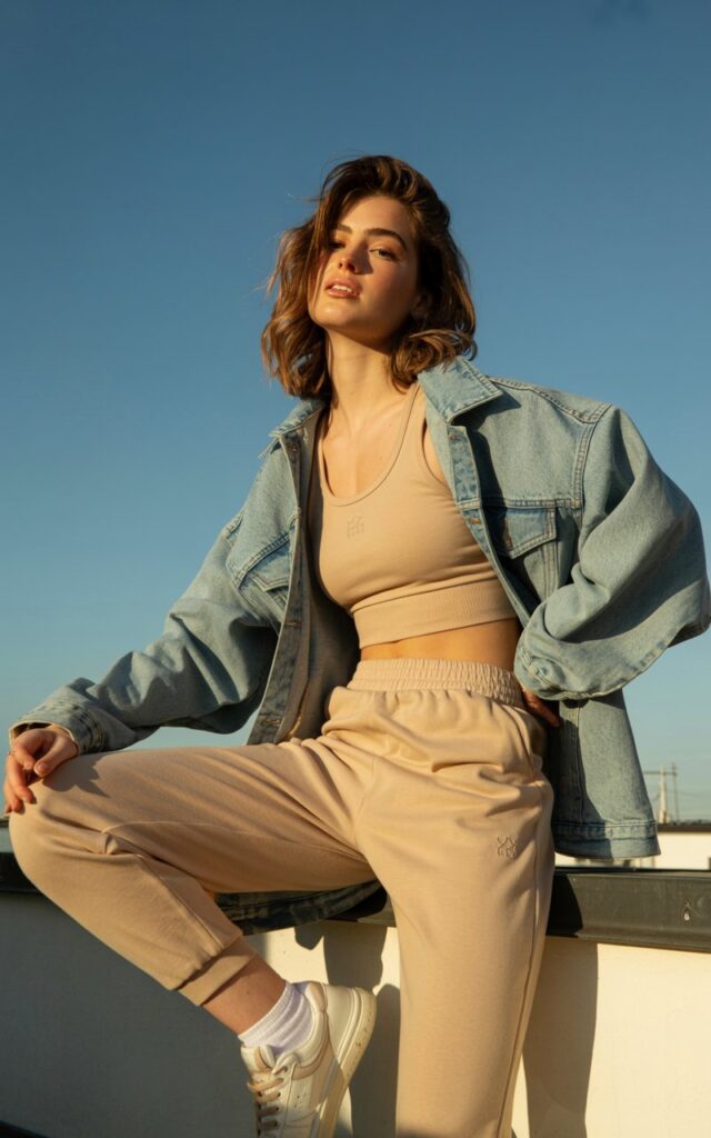 Sunny rooftop photoshoot. Model in a beige monochrome activewear set layered with a light-wash oversized denim jacket. Sneakers, tousled hair, and minimal makeup. Golden-hour light with clean, editorial framing.