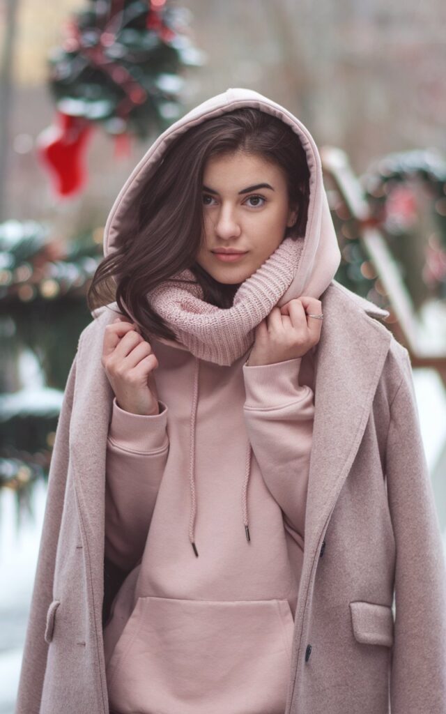 A natural-looking young woman wearing a blush hoodie layered under a classic wool coat. Theme pink christmas outfit