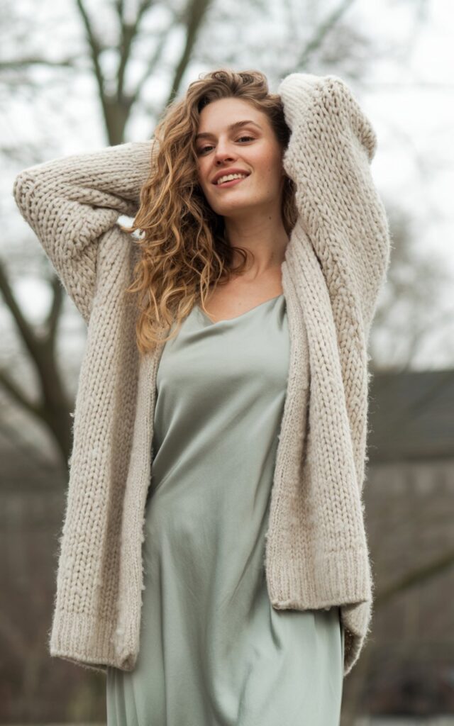 A natural-looking young woman wearing a chunky cardigan with a midi slip dress. Theme winter fashion outfits