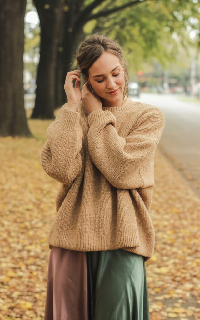 A natural-looking young woman wearing a chunky knit sweater layered over a satin midi skirt. Theme casual fall outfits