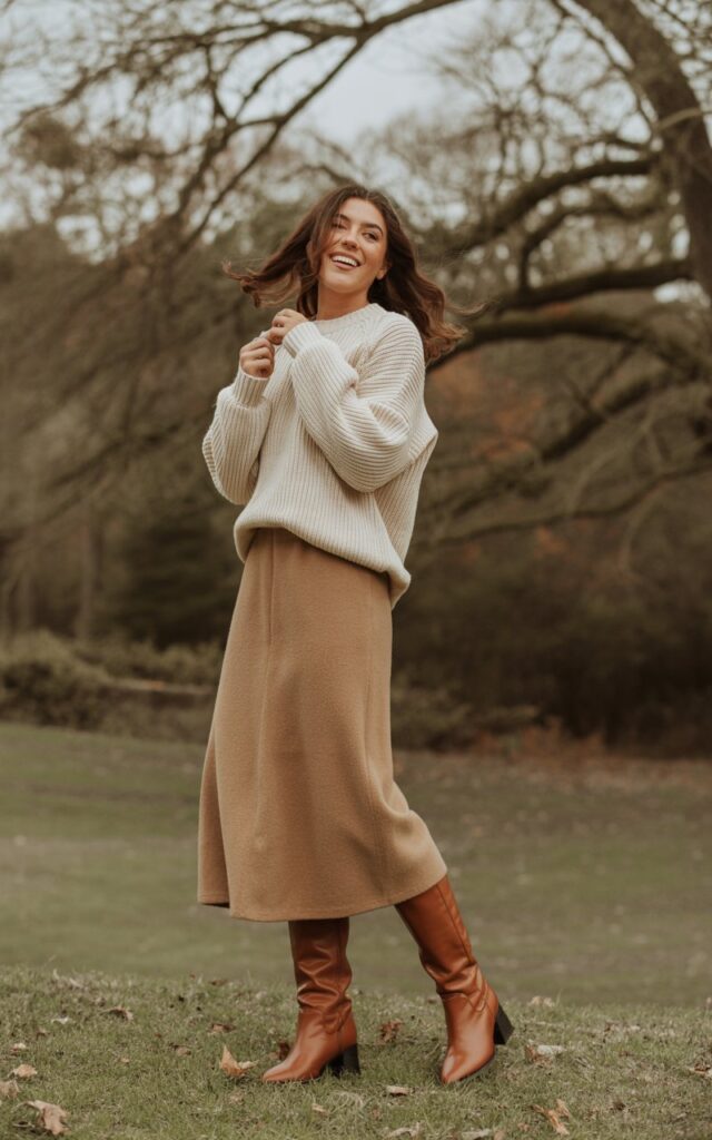A natural-looking young woman wearing a chunky sweater paired with a midi skirt and tall boots. Theme cold weather outfits