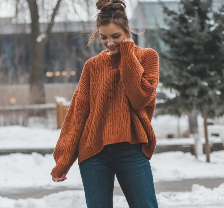 A natural-looking young woman wearing a cozy knit sweater layered over straight-leg jeans with ankle boots. Theme cold weather outfits